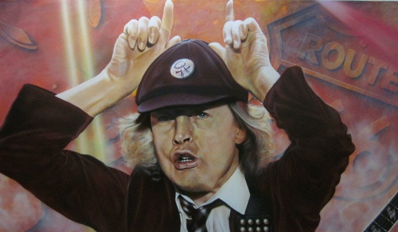 Angus Young is pictured holding his hands over his head in a horn motion. He is wearing a maroon suit and baseball hat, as well as a patterned tie. The background is a rust color with a sign reading Route 66.  