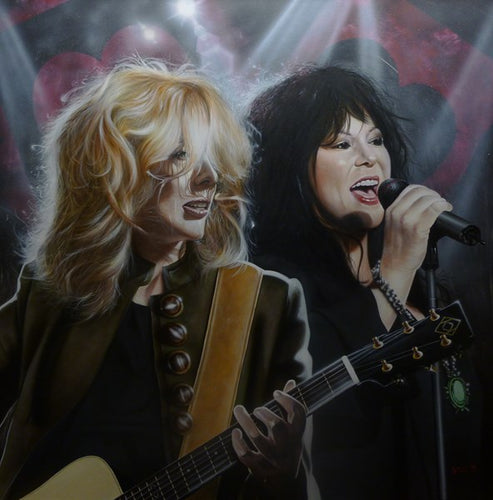Ann and Nancy Wilson are pictured on stage. Nancy Wilson is playing guitar in a buttoned green jacket and a black undershirt. Ann is singing into a microphone with a black jacket. Lights flash in a background with black and red hearts.