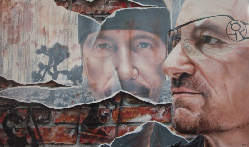 "Bono" from U2 is pictured on the right side, facing left, wearing glasses and a blank stare. In the background are bricks with black spraypaint showing the other members of the band. Also in the back is a large broken mirror with "The Edge's" face in it, wearing a beanie.